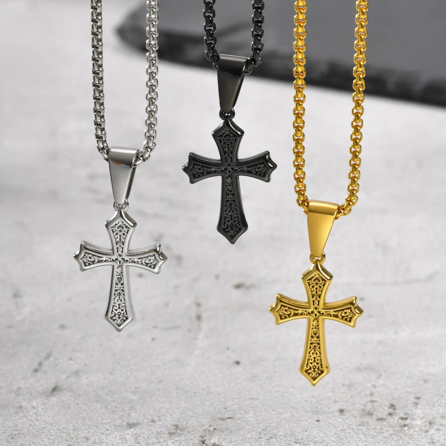 Trio of stainless steel cross pendant necklaces in silver, black, and gold with intricate gothic detailing, displayed on matching box chains for men.