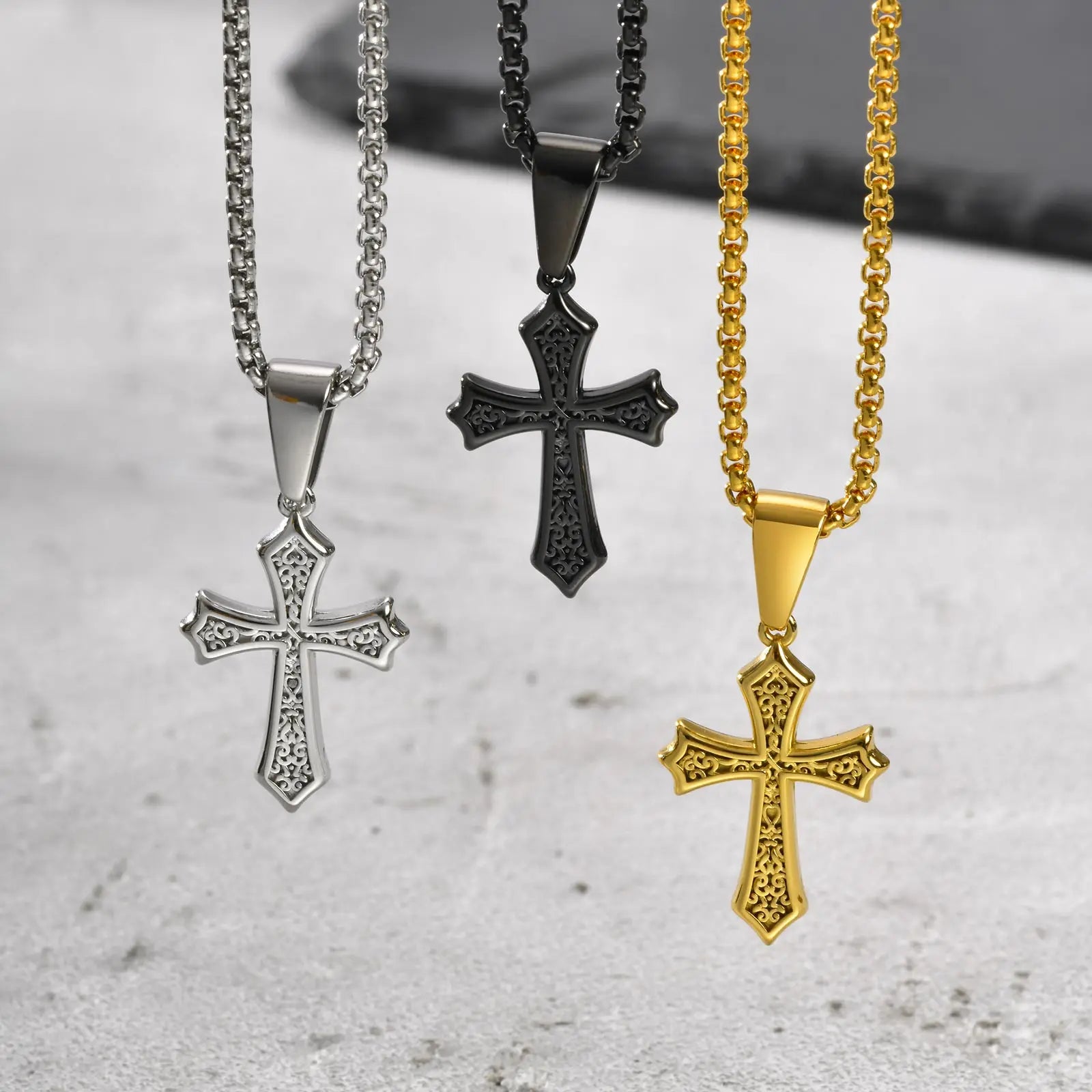 Trio of stainless steel cross pendant necklaces in silver, black, and gold with intricate gothic detailing, displayed on matching box chains for men.